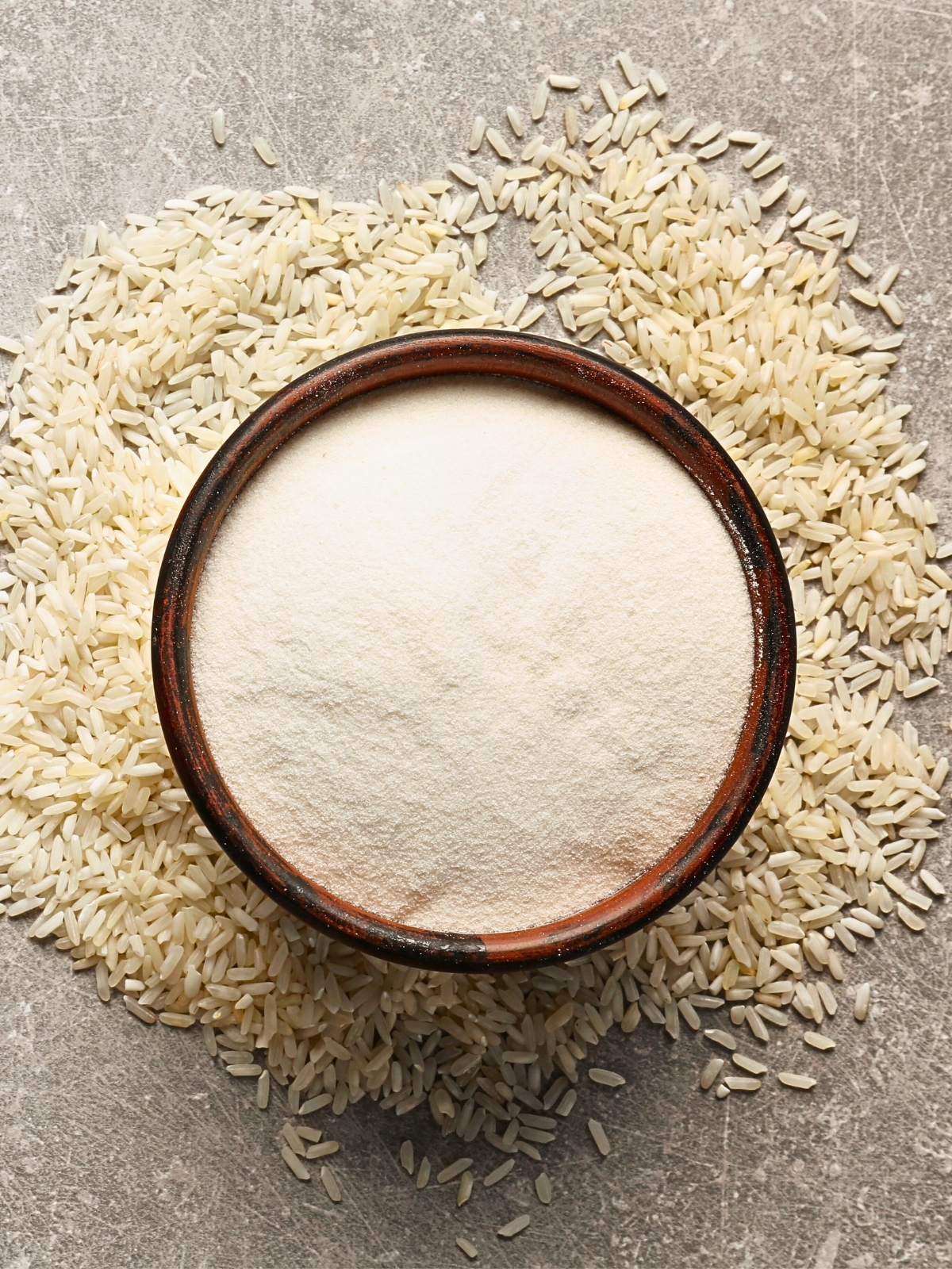 A bowl with Rice Flour and white rice grains around it on Gray Background